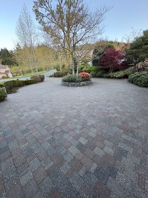 A wide view of a clean, professionally sealed multitone stone paver driveway featuring a circular garden bed and mature landscaping.