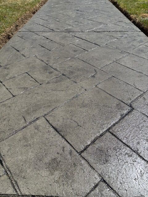 A close-up view of freshly sealed dark grey stamped concrete with a slate-pattern, showcasing a clean satin finish and weather protection.